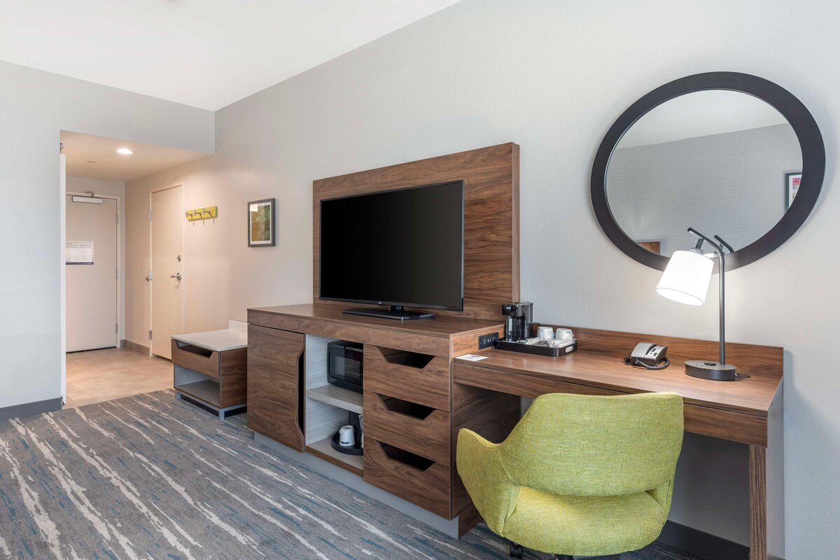 Ein Hotelzimmer mit einem Holztisch, einem Fernseher, einem Spiegel, einem grünen Sessel und Teppichboden.