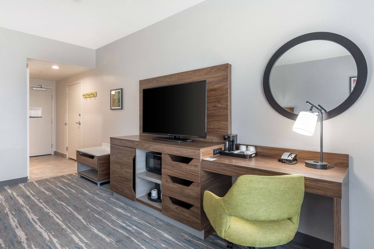A modern hotel room with a wooden desk, a television, a mirror, a green chair, and a carpeted floor.