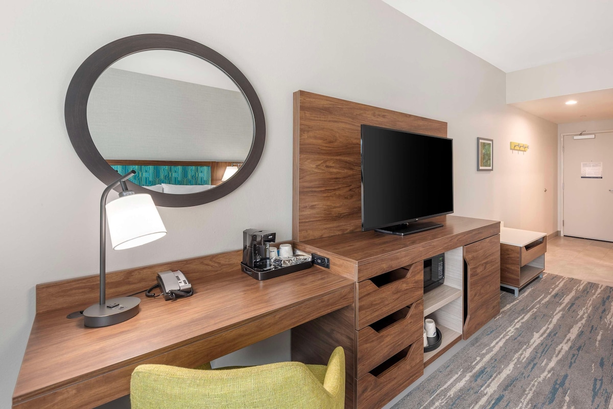 A hotel room with a wooden desk, a flat-screen TV, a mirror, a lamp, a telephone, and a green chair.