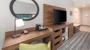 Ein modernes Hotelzimmer mit Schreibtisch aus Holz, einem Flachbildfernseher, einem runden Spiegel, einer Lampe, einem Telefon und einem grünen Sessel.