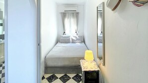 Een smalle gang die leidt naar een bed met een geruite vloer, een nachtkastje met een lamp en een spiegel aan de muur.