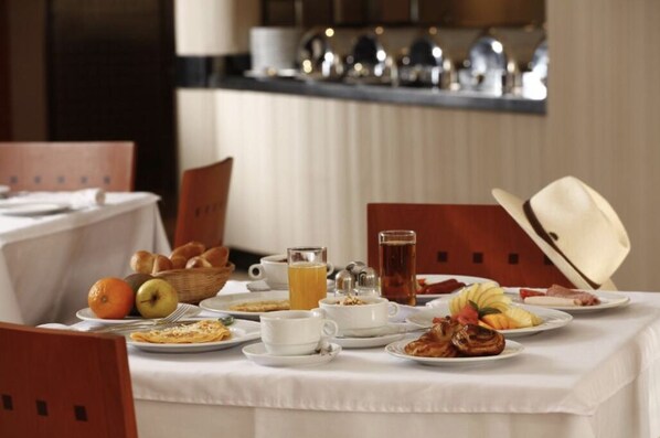 Une table dressée pour le petit-déjeuner, avec une variété de plats, notamment des fruits, des viennoiseries et des boissons, et recouverte d’une nappe blanche.