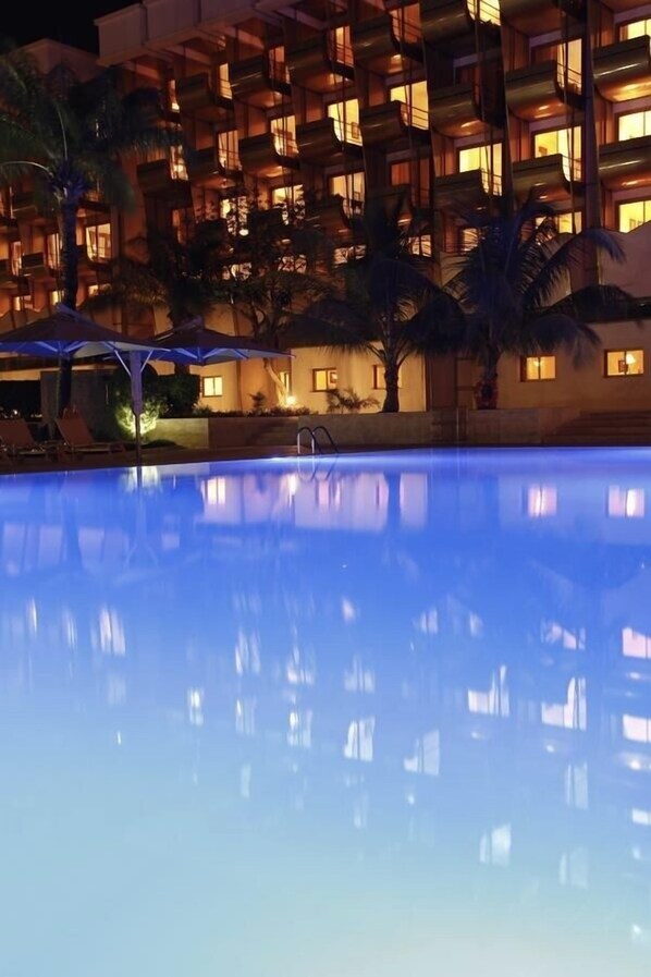 Une vue nocturne d’un hôtel avec une piscine, des palmiers et des balcons illuminés.