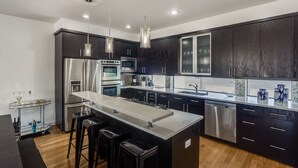 A modern kitchen with dark cabinetry, stainless steel appliances, a central island with stools, pendant lighting, and a large window. - Modern 4 Story Townhome -5 minutes walk to Deep Ellum & Fair Park! (Dallas)