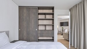 A modern bedroom with a large bed, a built-in wardrobe with multiple shelves, and a view into a living area with a TV and seating. - MOOI Apartments Grenchen (Grenchen)