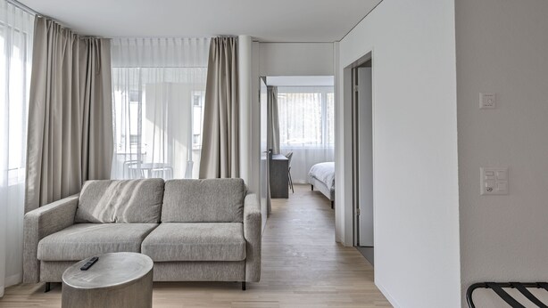 A modern living room with a grey sofa, a round coffee table, and a bed in the background. - MOOI Apartments Grenchen (Grenchen)