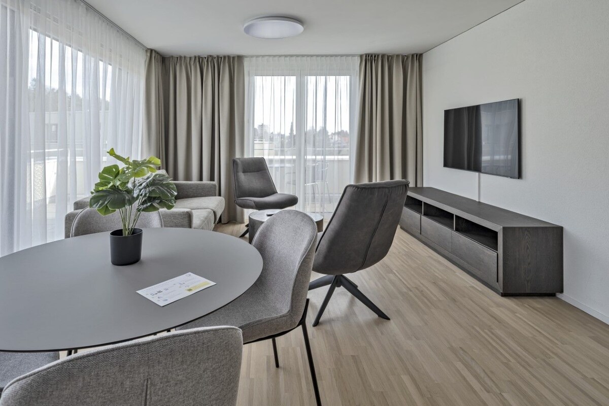 A modern hotel room with a round table, grey chairs, a grey sofa, a TV mounted on the wall, and a large window with curtains.