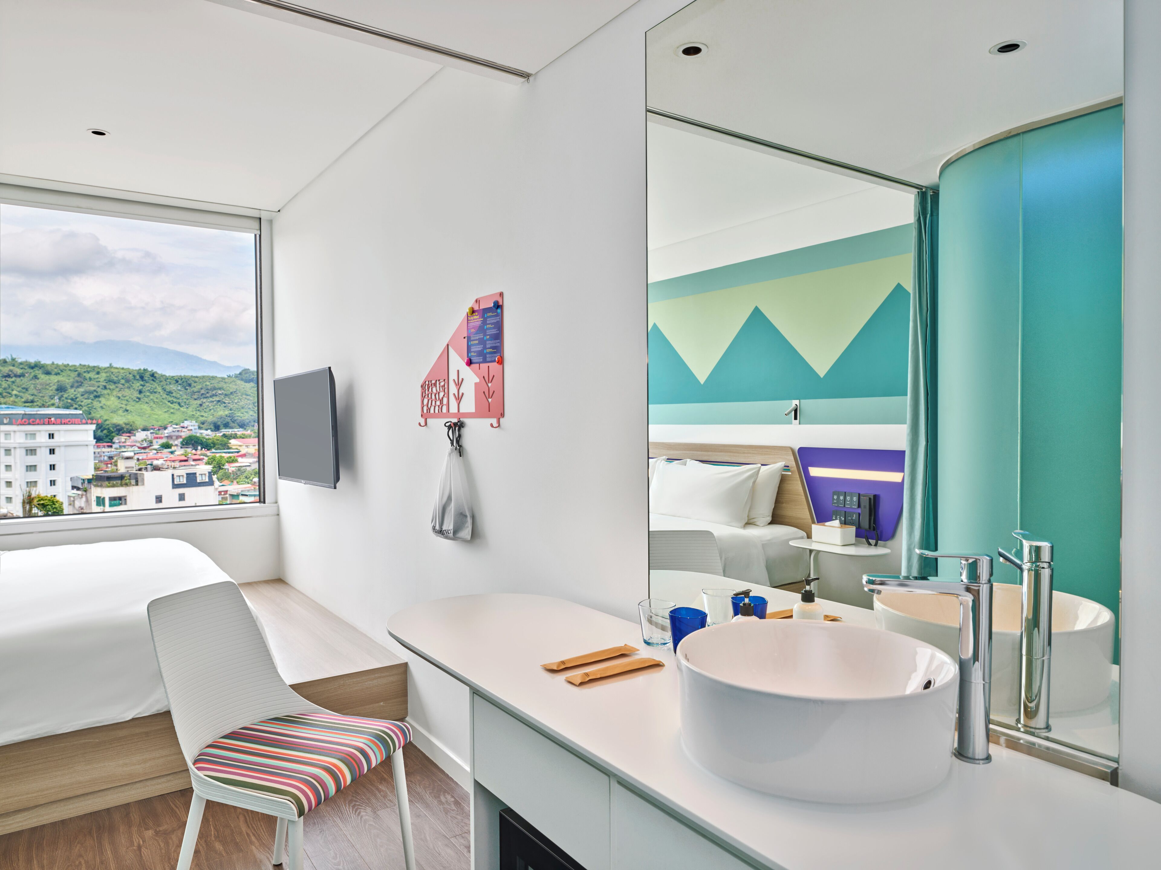 A modern hotel room with a bed, a desk, a large mirror, a bathroom with a sink, and a view of the city.