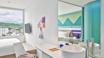 Une chambre d’hôtel moderne avec un grand lit, un bureau, une chaise agrémentée d’un coussin coloré, un grand miroir, un lavabo blanc et une vue sur la ville par la fenêtre.