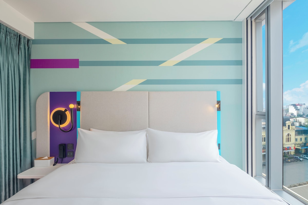 A modern hotel room with a large bed, a headboard with a circular light, and a city view through the window.