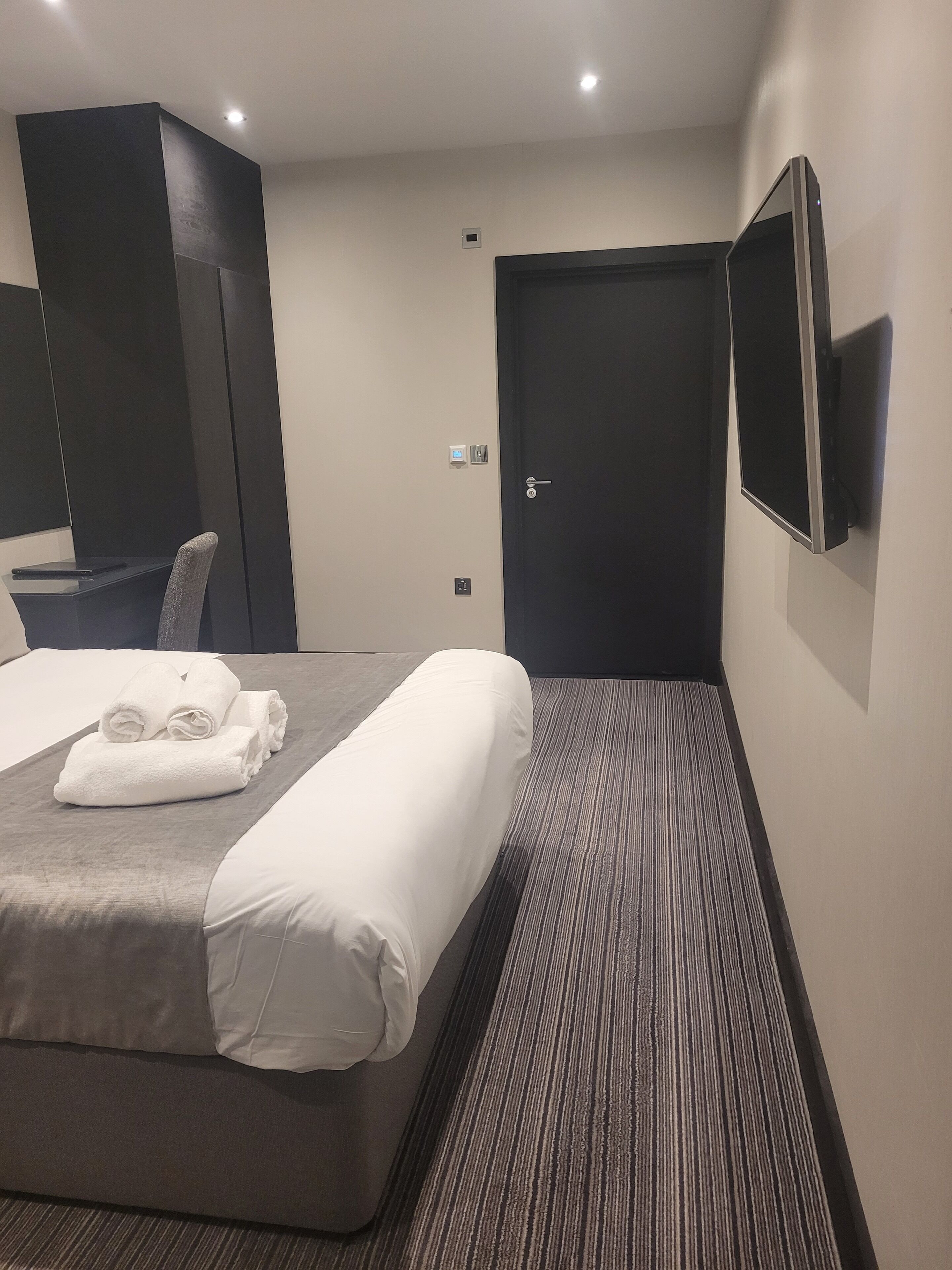 A hotel room with a bed, a desk, a chair, a flat-screen TV, and a door.