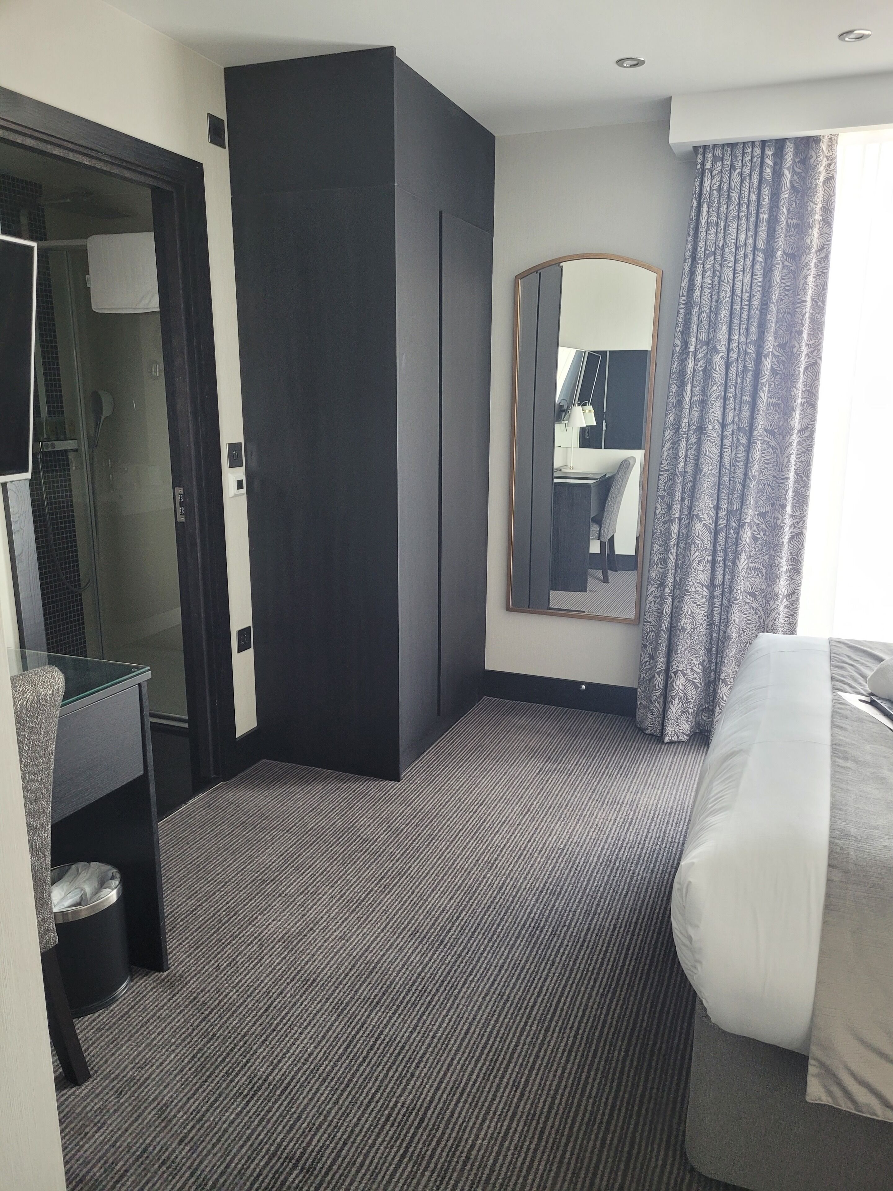 A hotel room with a bed, a desk, a mirror, a wardrobe, and a bathroom visible through an open door.