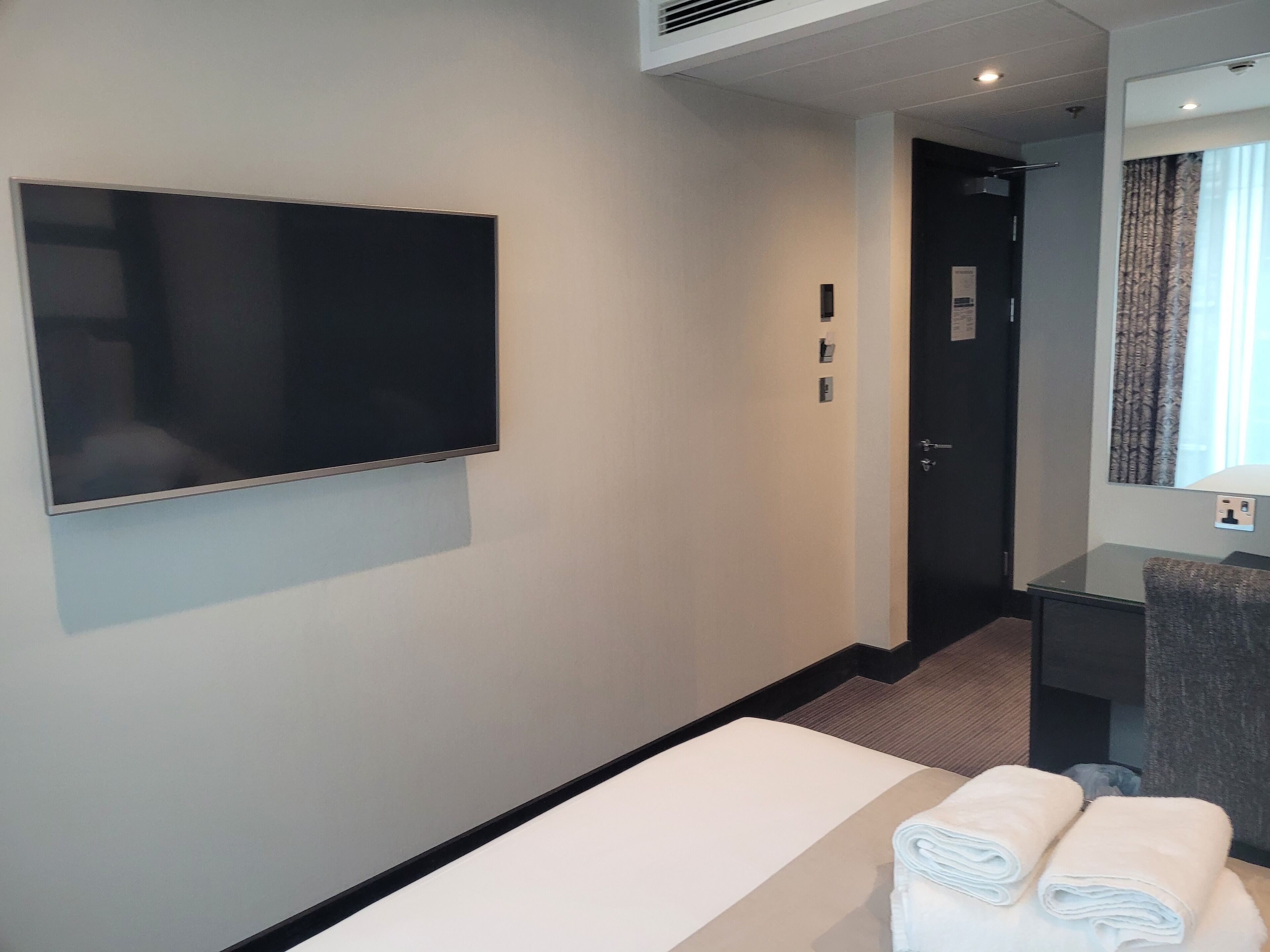 A hotel room with a large flat-screen TV mounted on the wall, a bed with white linens, a small desk with a mirror, and a door leading to a bathroom.
