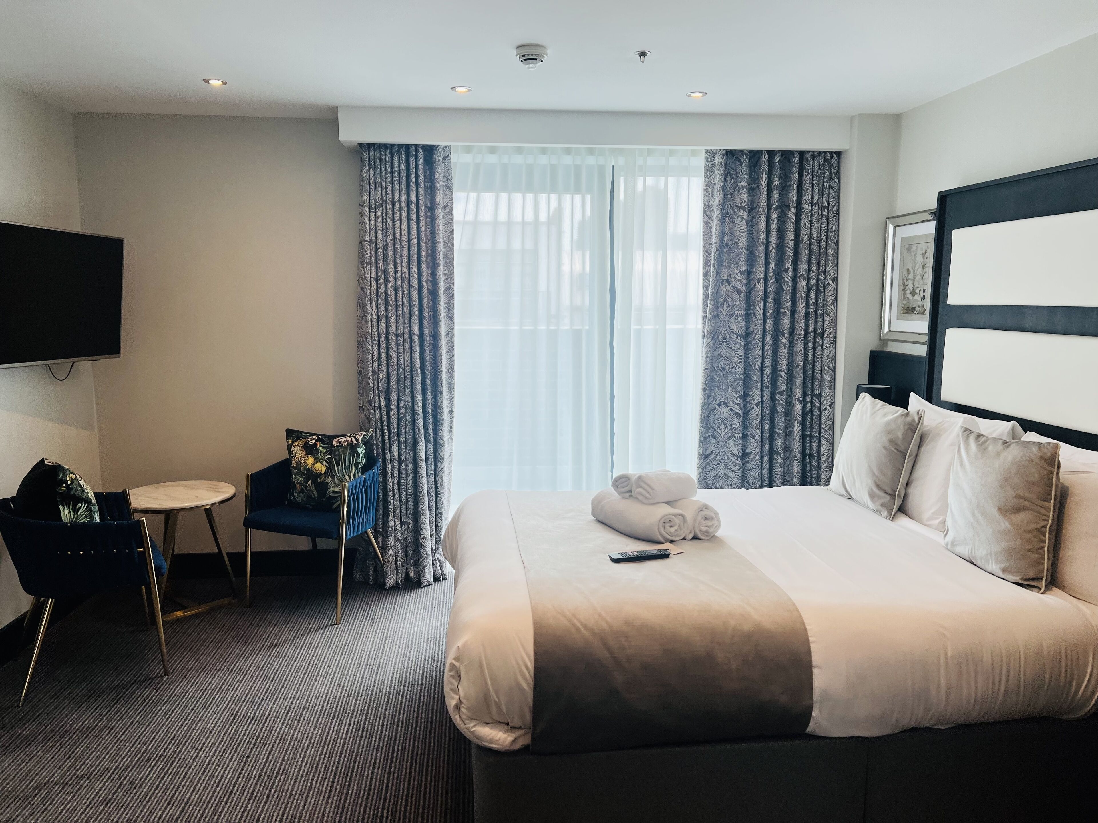 A hotel room with a large bed, a small desk, a chair, a TV, and a window with curtains.