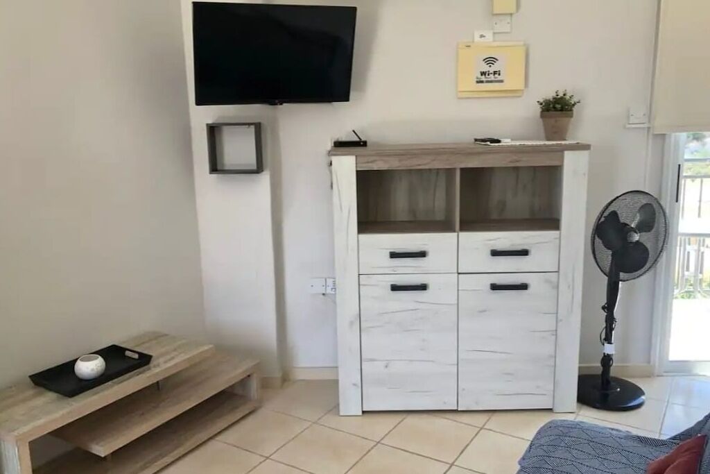 Ett modernt vardagsrum med en platt-TV som är monterad på väggen, ett vitt skåp med lådor, ett litet bord med en dekorativ prydnad och en golvfläkt.