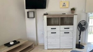 A modern living room with a flat-screen TV mounted on the wall, a white cabinet with drawers, a small table with a decorative item, and a standing fan.