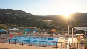 A swimming pool with lounge chairs and umbrellas, surrounded by a fence, with a mountainous landscape in the background.