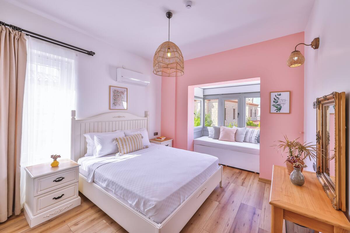 A bedroom with a bed, bedside tables, a window seat, a mirror, and a view of the outside.
