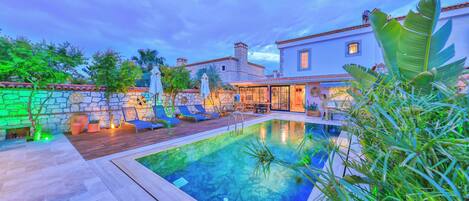 A pool area with a stone wall, lounge chairs, and a building with a tiled roof.