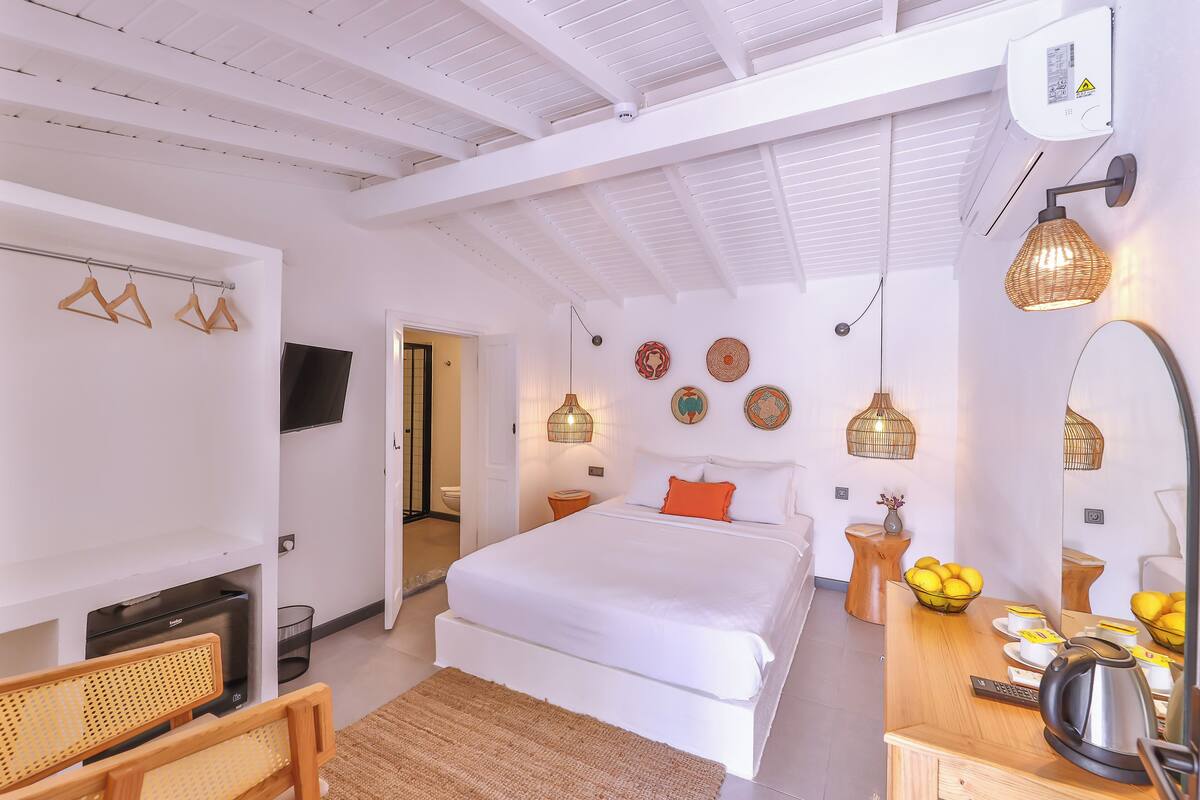 A modern bedroom with a large bed, a desk with a kettle, and a bathroom visible through an open door.