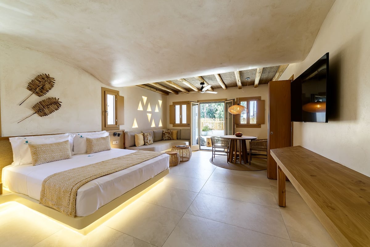 A bedroom with a large bed, a wooden bench, a dining table, and a TV mounted on the wall.