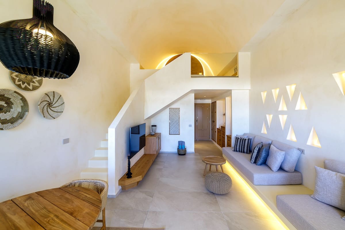A modern interior with a wooden dining table, a sofa with cushions, a staircase, and decorative wall lights.