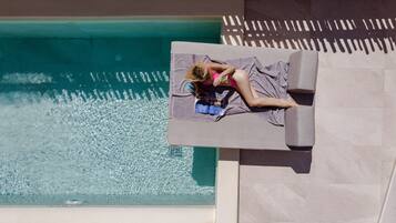 Un área de descanso junto a la piscina con una persona relajándose en un banco acolchado, un libro y un par de gafas de sol.