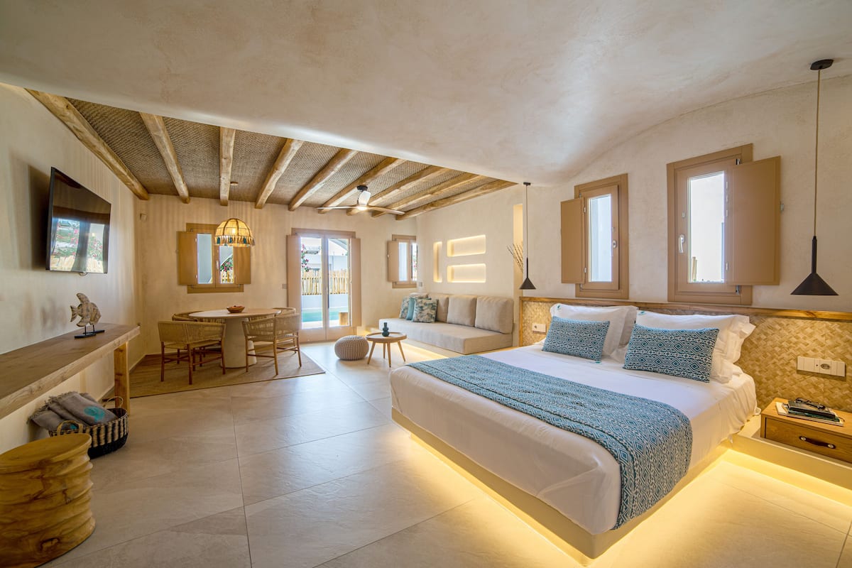 A spacious bedroom with a large bed, a wooden ceiling, a dining area with a table and chairs, and a view of the outdoors through a door.