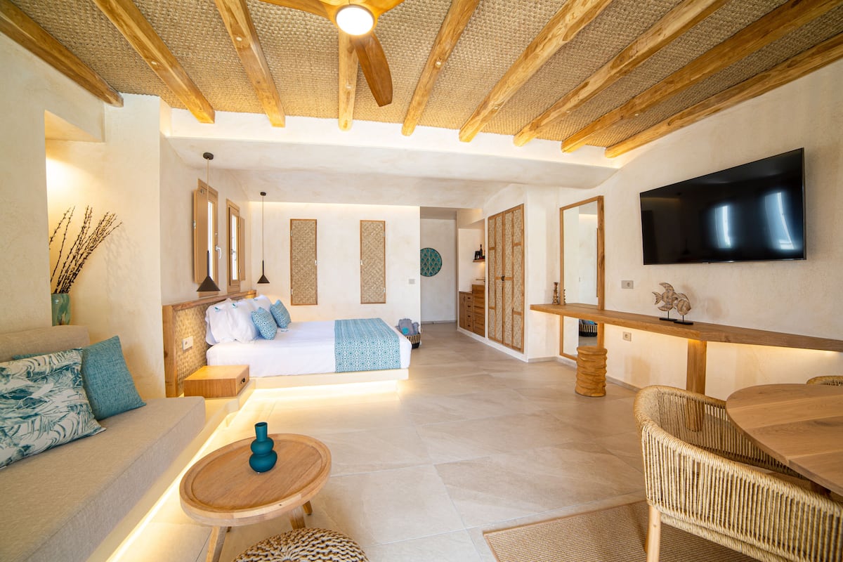 A modern hotel room with a large bed, a sitting area with a sofa and a small table, a dining area with a wooden table and chairs, and a TV mounted on the wall.