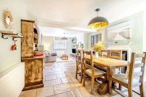A dining area with a wooden table and chairs, a hanging pendant light, and a kitchen area with a wooden cabinet and shelves.
