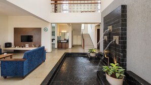 A spacious living area with a blue sofa, a flat-screen TV, a kitchen in the background, and a black tiled wall with a water feature. - CosmicStays GreyStone Villa Mountain View & Dam (Paud)