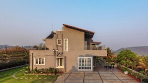 A modern two-story house with a balcony, a small garden, and a paved patio area. - CosmicStays GreyStone Villa Mountain View & Dam (Paud)