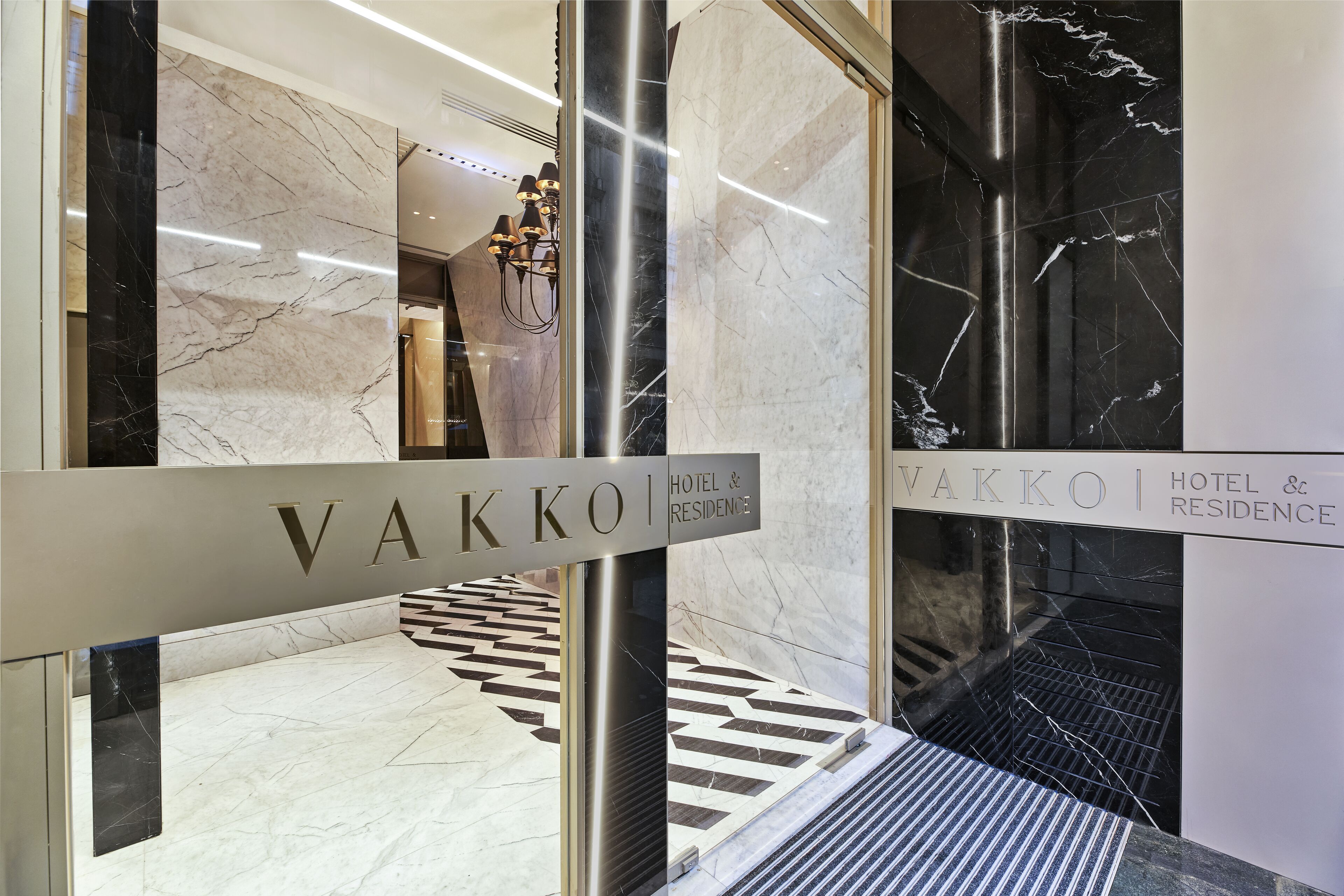 A modern hotel lobby with marble flooring, a black and white patterned area rug, and a staircase leading to an upper level.