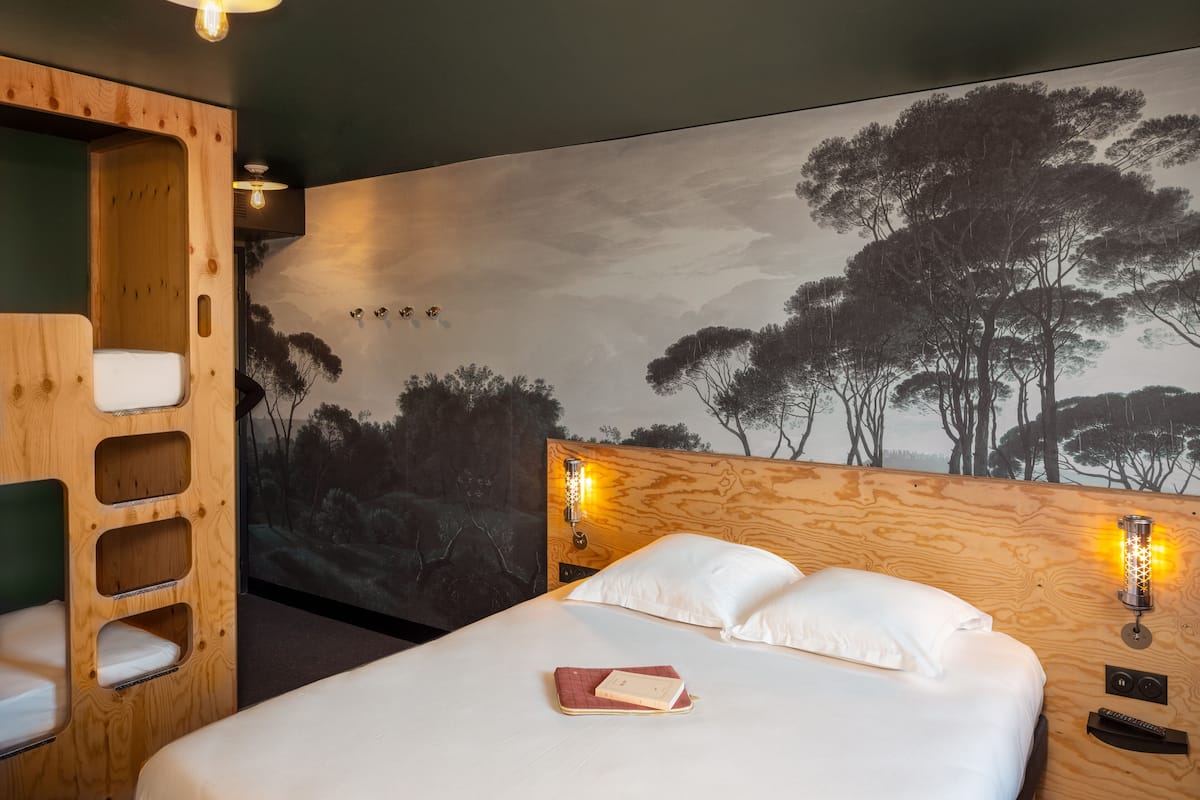 A bedroom with a bed, a wooden headboard, a mural of trees, and two wall-mounted lights.