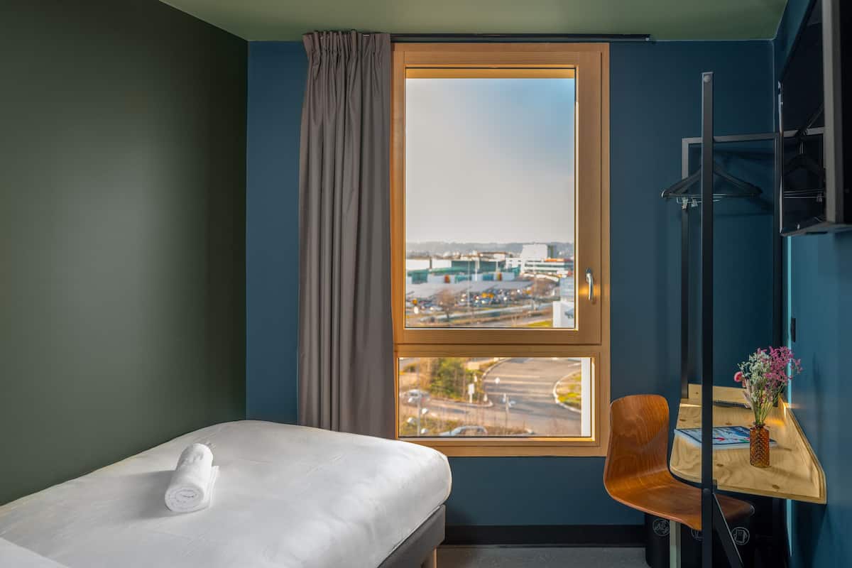 A hotel room with a bed, a chair, a small table with a vase, and a window with a view of an airport.