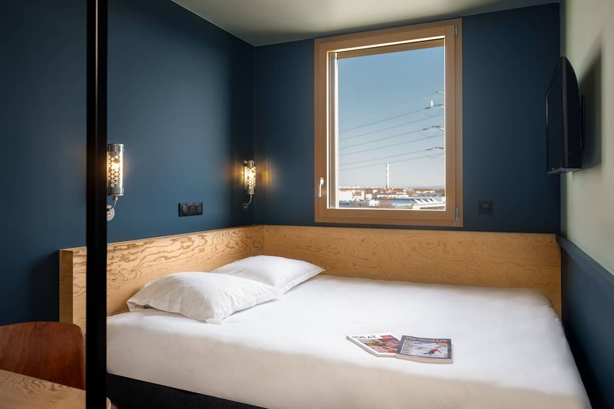 A hotel room with a bed, two pillows, a magazine, a window with a view of buildings, and a wall-mounted TV.
