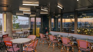 Un espacio para comer al aire libre con sillas y mesas de mimbre, amplias ventanas con vistas a la ciudad e iluminación moderna.