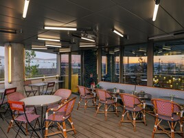 Uma área de refeições ao ar livre com cadeiras e mesas de vime, grandes janelas com vista para a cidade e luminárias modernas.