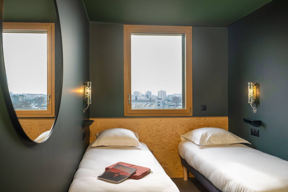A hotel room with two beds, large windows, and wall-mounted light fixtures.