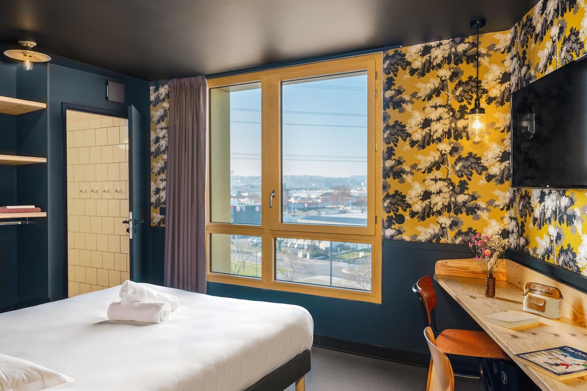 A hotel room with a bed, a desk, a chair, a TV, and a window with a city view.