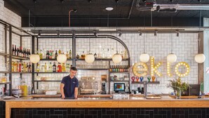 Un bar con un barman, estantes llenos de licores, una máquina de café y un letrero de neón.