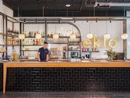 Un bar con un barman, estantes llenos de licores, una máquina de café y un letrero de neón.