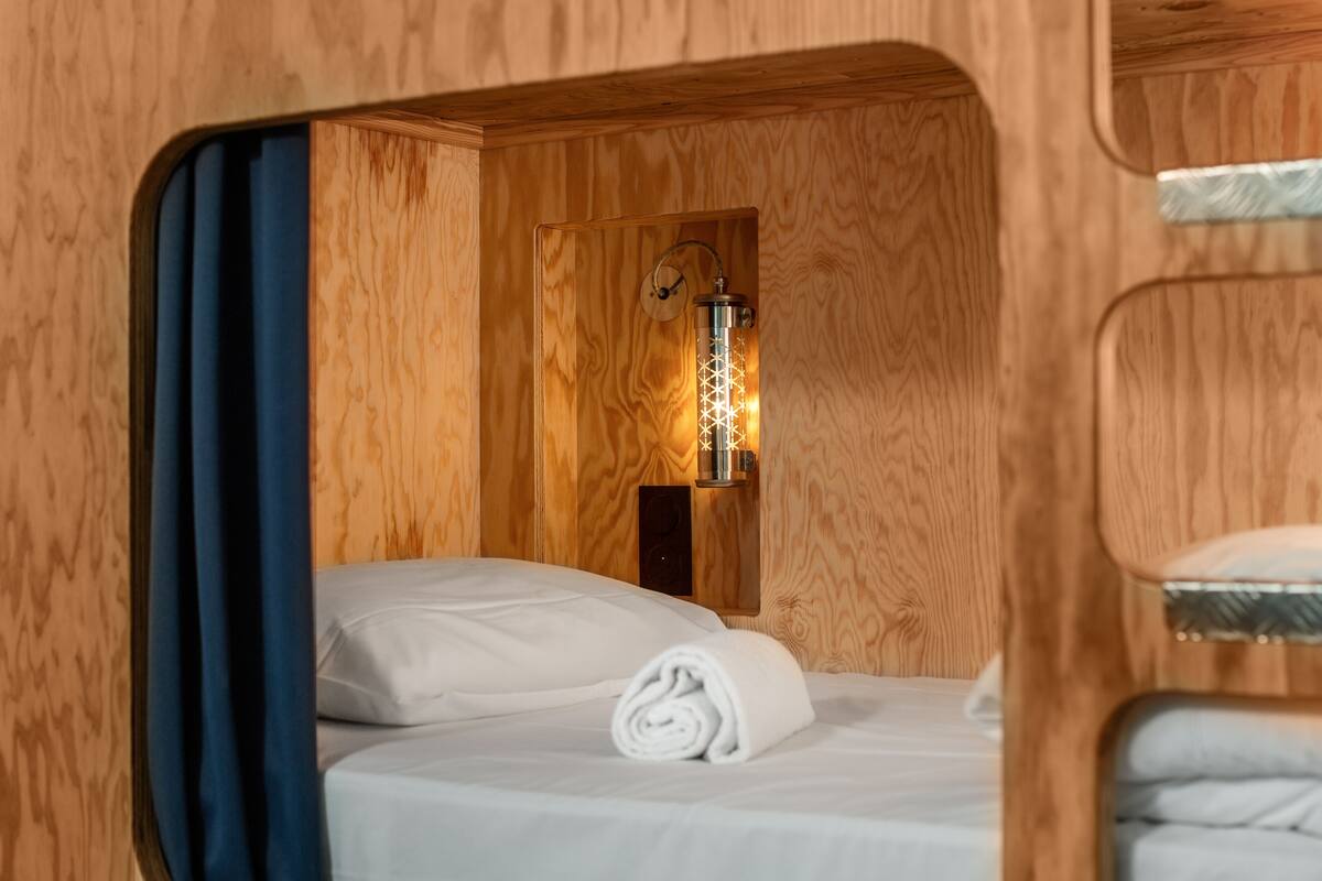 A compact wooden room with a bed, a wall-mounted light fixture, and a window.