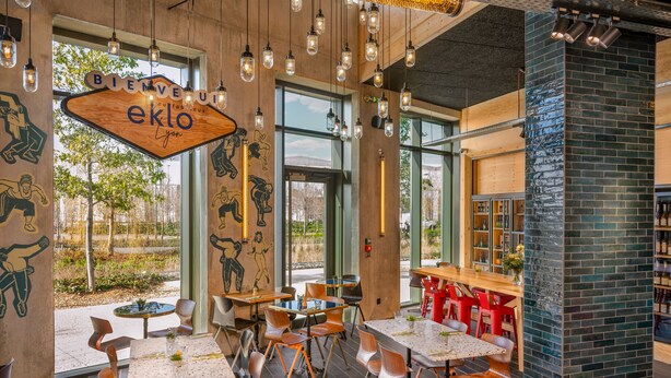 Un interior de restaurante moderno con mesas y sillas de madera, ventanas amplias y una pared decorativa de ladrillo.