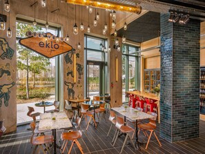 A modern restaurant interior with wooden tables and chairs, large windows, and a brick accent wall. - Eklo Lyon Sud Eurexpo (Venissieux)