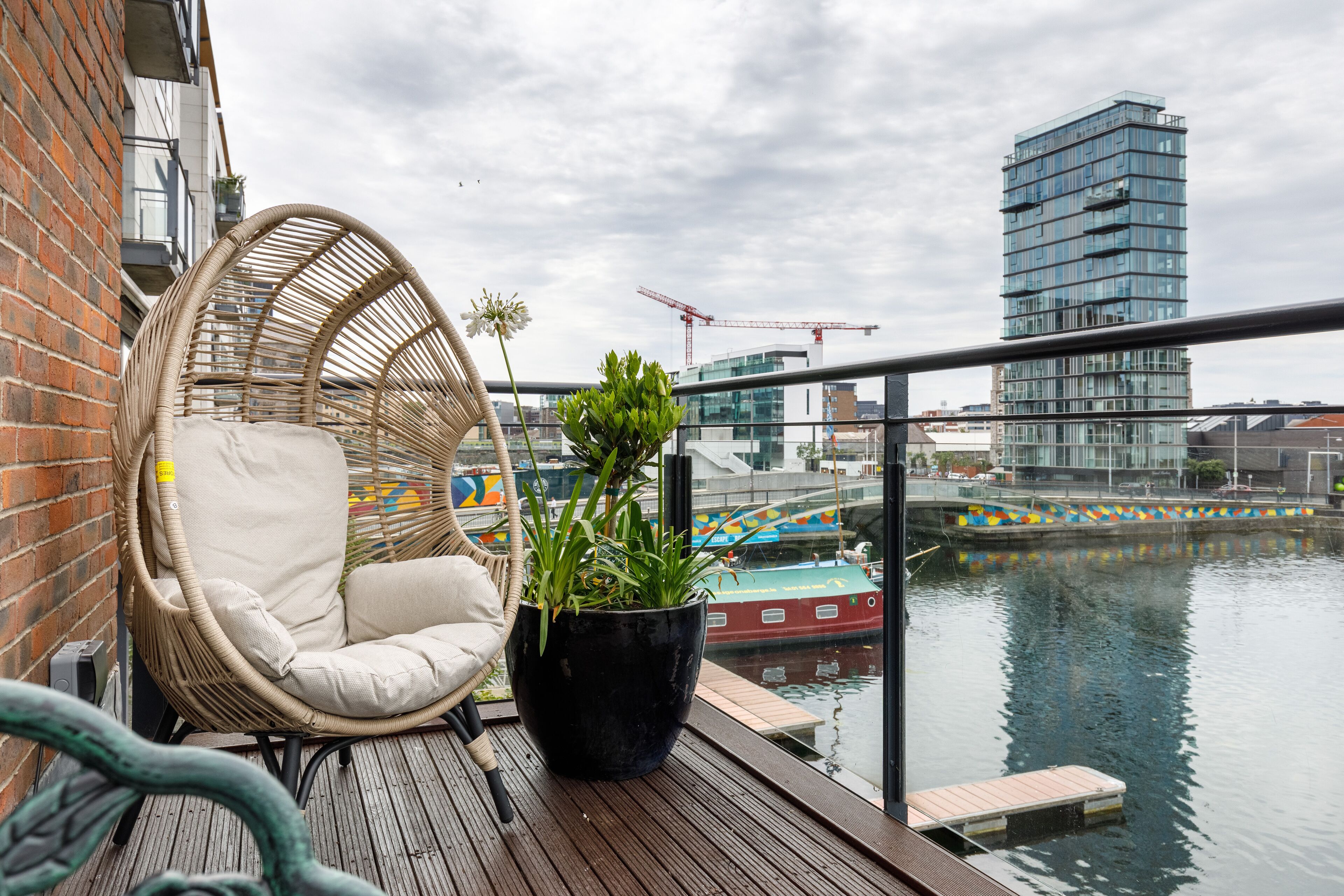 A wicker chair with a cushion and a potted plant on a wooden deck overlooking a body of water with a modern building and a bridge in the background.