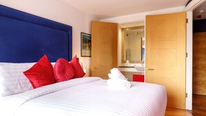 Une chambre d’hôtel avec un grand lit, des oreillers rouges et blancs, une tête de lit bleue et une porte ouverte donnant sur une salle de bain équipée d’un miroir et d’un lavabo.