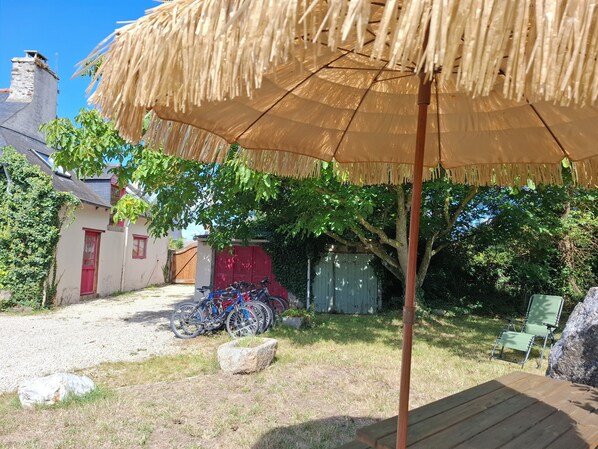 Eine Terrasse mit einem Reetdachschirm, Fahrrädern, einem Holztisch und einem Garten mit Bäumen und einem Schuppen.