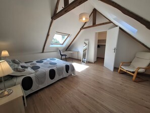 Ein Schlafzimmer mit schrägen Wänden, einem Bett mit gemustertem Überwurf, einem Schreibtisch, einem Stuhl, einem Spiegel und einem Kleiderschrank.