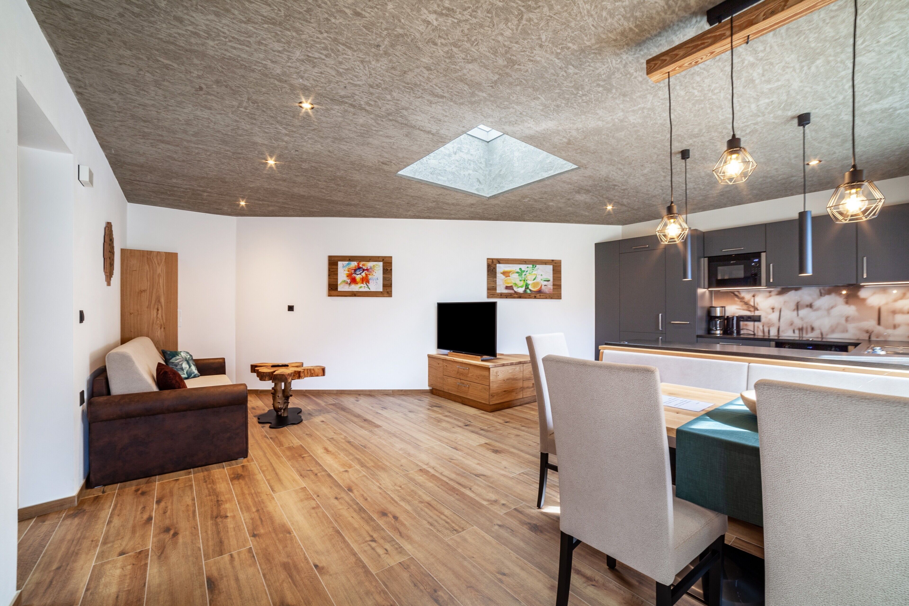 A modern living space with a wooden floor, a sofa, a dining table with chairs, a TV, and kitchen cabinets.